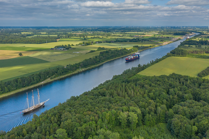 Ein Frachtschiff und ein Segelboot fahren auf dem Nord-Ostsee-Kanal durch grüne Landschaft bei Kiel.