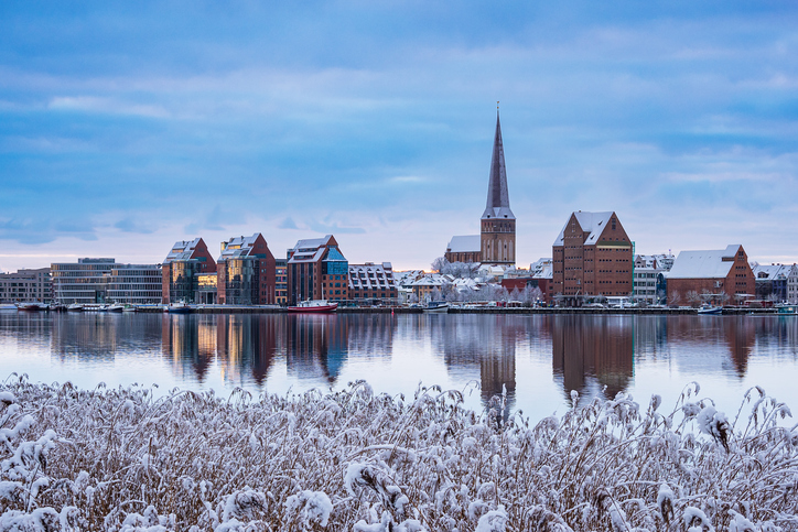 Blick über schneebedecktes Schilf auf die Silhouette der Stadt mit der Petrikirche, die sich im spiegelglatten Wasser der Warnow spiegelt.