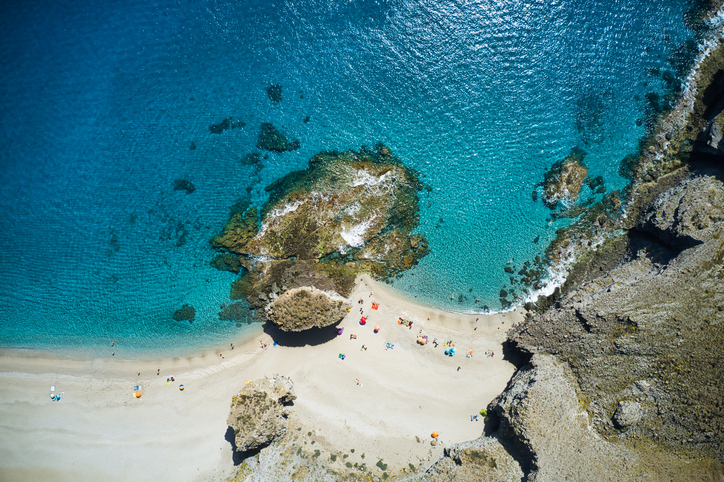 Senkrechter Blick von oben auf türkisfarbenes Wasser, einen hellen Sandstrand und markante Felsformationen im Meer.