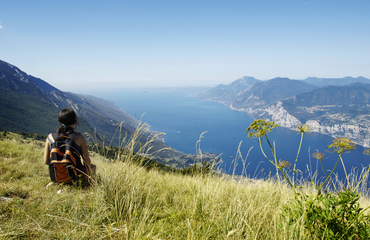 Wanderin mit Rucksack genießt den Panoramablick auf den tiefblauen Gardasee von einem Berggipfel aus.