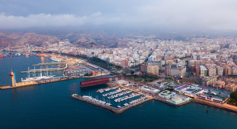 Panorama des Hafens von Almería bei Dämmerung mit beleuchteten Stegen, Segelbooten und der Stadt im Hintergrund.