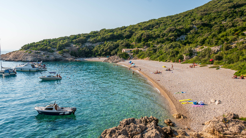 Ein Blick auf einen Kiesstrand auf der Insel Cres mit türkisfarbenem Wasser und kleinen Booten in der Bucht.