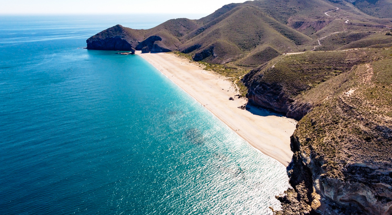 Ein weiter, heller Sandstrand schmiegt sich an eine tiefblaue Bucht, umgeben von trockenen, vulkanischen Hügeln.