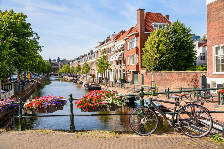 Ein schwarzes Fahrrad lehnt an einem Brückengeländer mit leuchtend roten und lila Blumenkästen über einer malerischen Gracht.