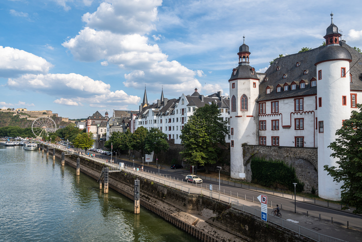 Blick entlang des Rheinufers auf das weiße Kurfürstliche Schloss mit seinen runden Türmen und einem Riesenrad in der Ferne.