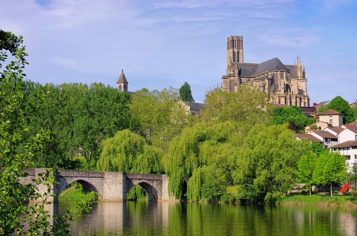 Die Kathedrale von Limoges ragt hinter Bäumen, Brücke und ruhigem Wasser in den Himmel.