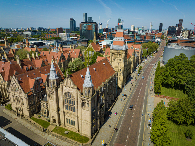 Luftaufnahme der neugotischen Universitätsgebäude mit roten Ziegeldächern, angrenzend an eine breite Straße und die moderne Skyline im Hintergrund.
