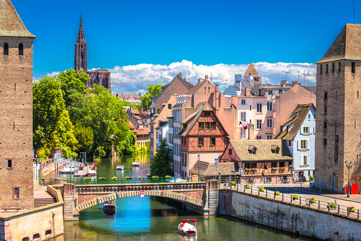 Blick entlang der Ill auf Fachwerkhäuser und eine Steinbrücke mit kleinen Elektrobooten auf dem Wasser.