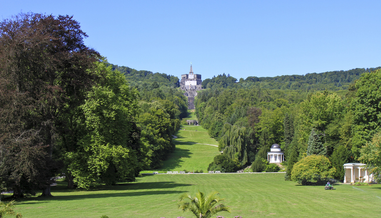 Eine weite Sichtachse über eine grüne Wiese direkt hinauf zum Herkules-Monument, flankiert von dichtem Wald und kleinen Tempeln.