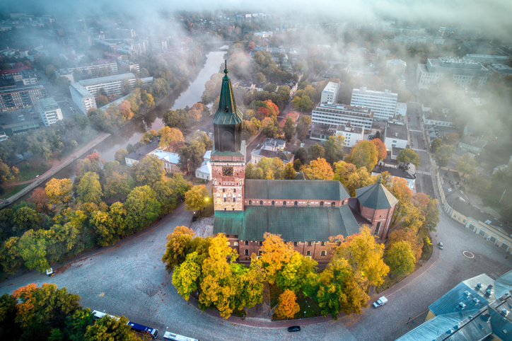 Drohnenaufnahme des Doms von Turku im Herbst, umgeben von bunt gefärbten Bäumen und leichtem Nebel über dem Aura-Fluss.