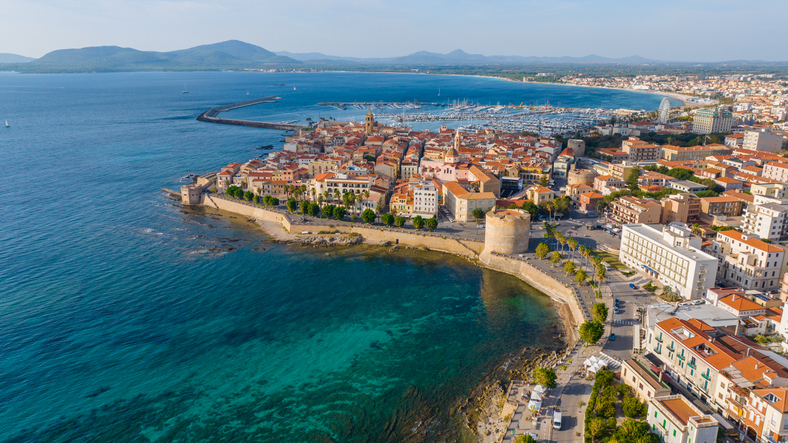 Panorama-Luftbild von Alghero auf Sardinien mit Stadtmauer, Yachthafen und dem türkisfarbenen Mittelmeer