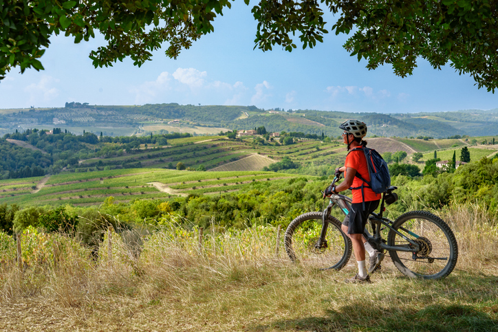 Ein Radfahrer hält mit seinem Mountainbike an einem Aussichtspunkt und blickt über die hügeligen Weinreben der Toskana.