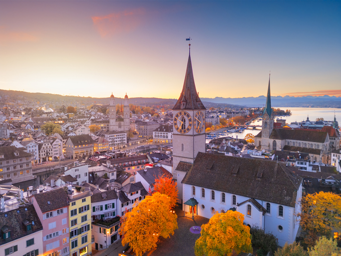 Das beleuchtete Zifferblatt der St. Peter Kirche während der Dämmerung mit Blick über die Dächer der Altstadt.