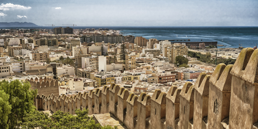 Sicht durch die gezackten Zinnen einer Steinmauer auf das dichte Häusermeer von Almería und das nahegelegene Mittelmeer.