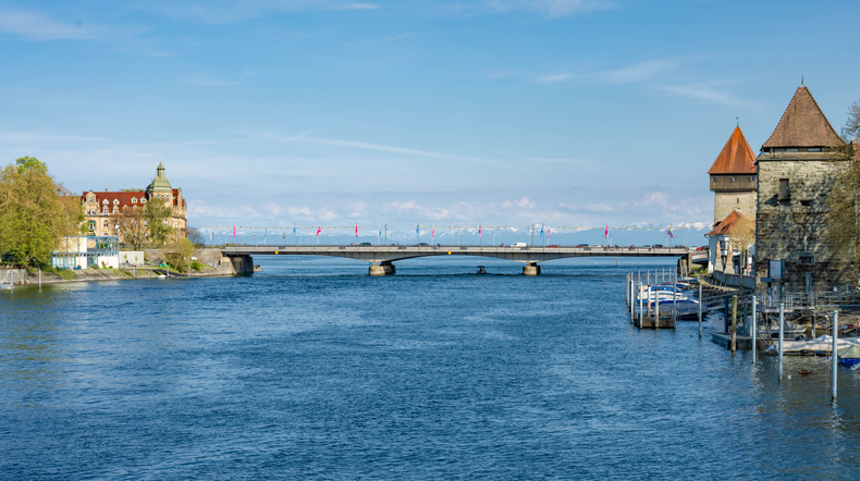 Blick vom Wasser auf die Rheinbrücke in Konstanz, flankiert von historischen Türmen und modernen Hotelbauten.