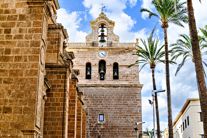 Nahaufnahme des Glockenturms der Kathedrale von Almería mit einer Uhr und sichtbaren Glocken unter einem blau-weißen Himmel.