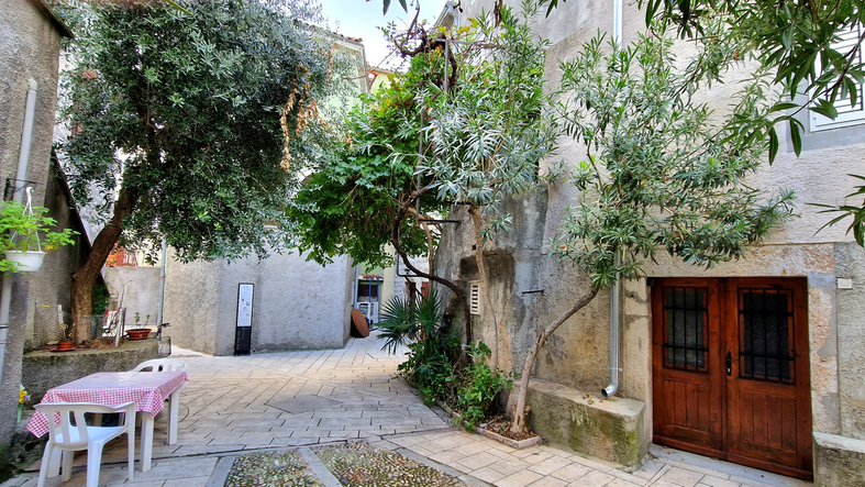 Eine schmale, schattige Gasse auf Cres mit alten Steinhäusern, Olivenbäumen und einem gedeckten Tisch im Freien.