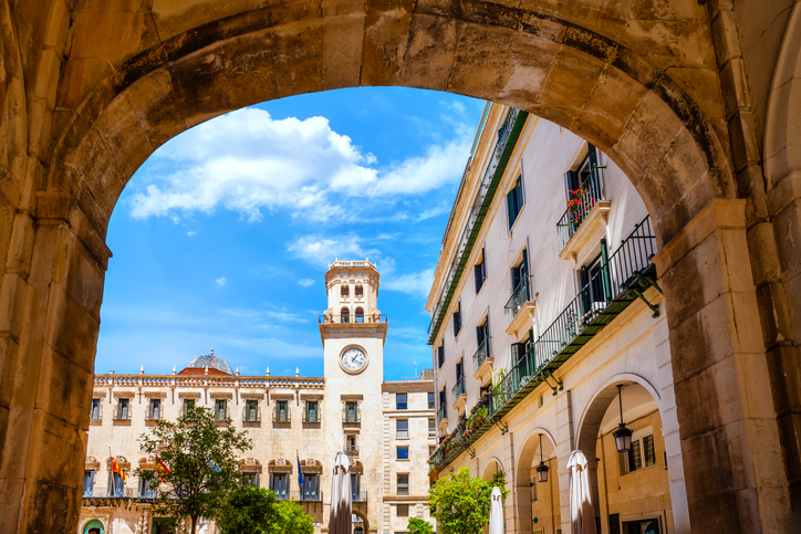 Der historische Rathausplatz in Alicante, gesehen durch einen steinernen Torbogen mit blauem Himmel