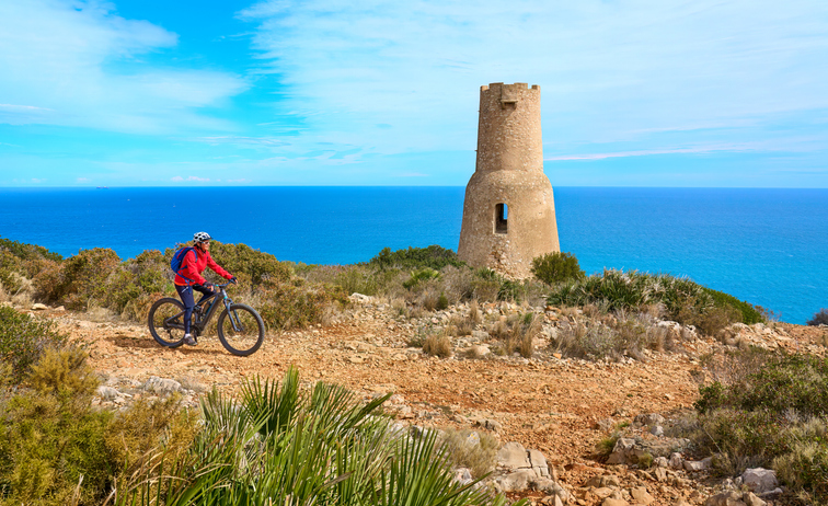 Ein Radfahrer fährt auf einem steinigen Pfad an der Küste entlang, im Hintergrund steht ein massiver, runder Wachturm aus Stein vor dem blauen Meer.