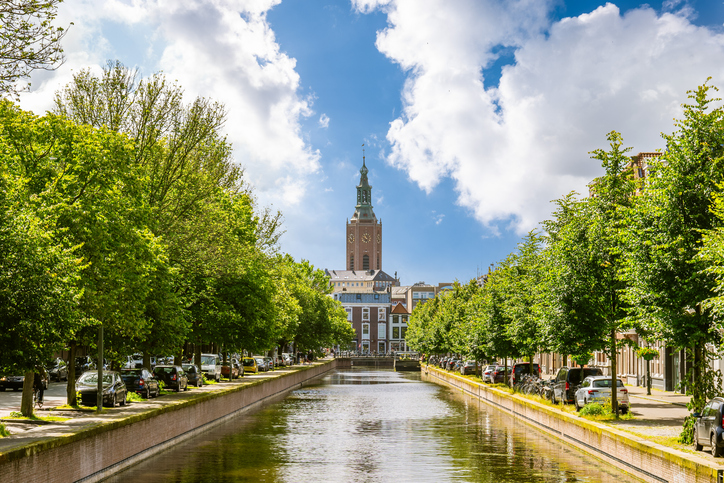 Ein ruhiger Kanal im Stadtzentrum von Den Haag, gesäumt von grünen Bäumen, mit dem hohen Turm der Grote Kerk im Hintergrund.