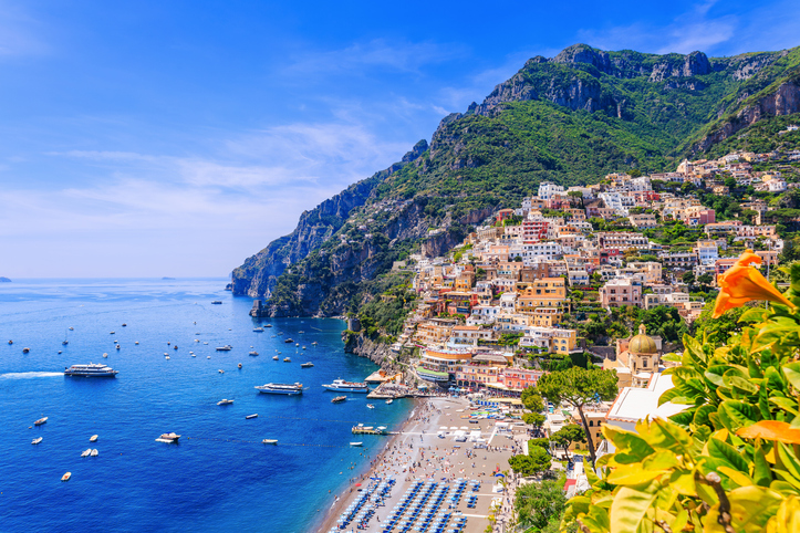 Ein sonniger Blick auf Positano von oben, mit vielen bunten Häusern am Hang, einem vollen Strand und blühenden Blumen im Vordergrund.