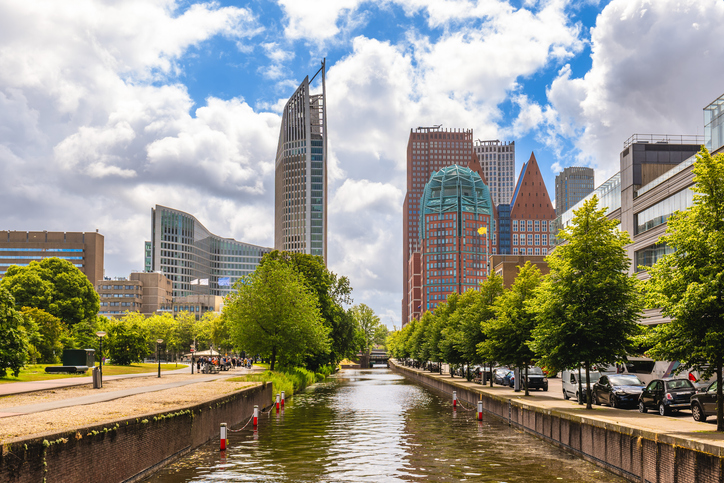 Ein von Bäumen gesäumter Kanal im Vordergrund, dahinter ragen die futuristischen Spitzen der Ministeriumsgebäude in den bewölkten Himmel.