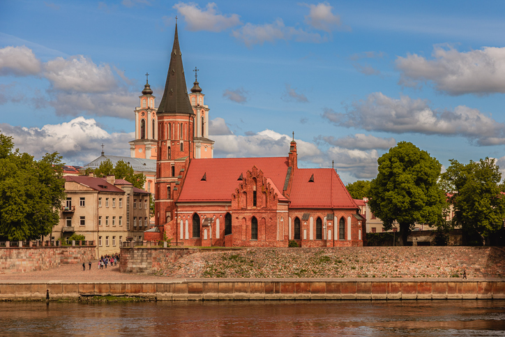 Eine rote Backsteinkirche mit spitzem Turm direkt am Flussufer, dahinter die Türme einer weiteren Barockkirche.