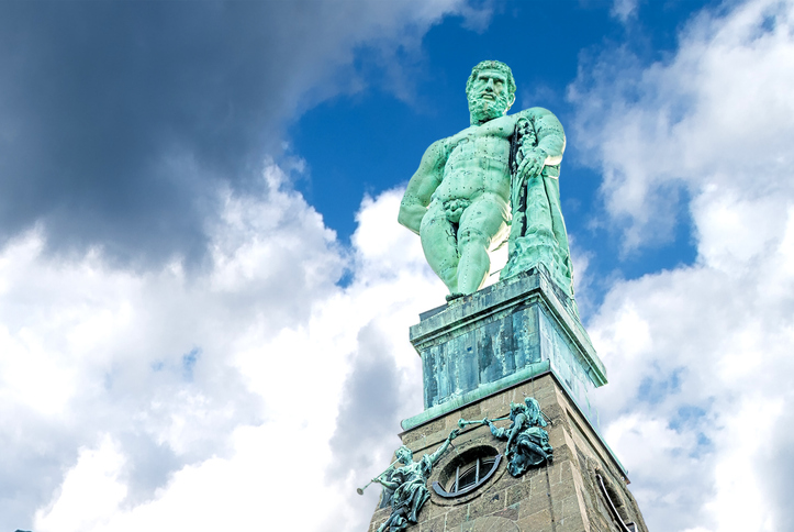 Eine detailreiche Ansicht der kupfergrünen Herkules-Statue auf ihrem Sockel gegen einen dramatisch bewölkten Himmel.