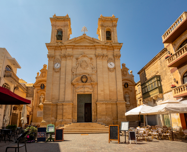Eine imposante barocke Kirche mit zwei Türmen steht an einem sonnigen Platz mit Straßencafés in der Hauptstadt von Gozo.
