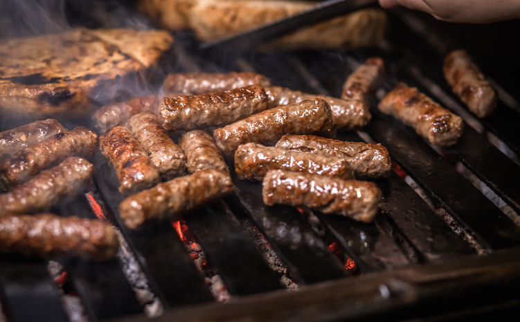 Saftige Cevapcici brutzeln auf einem heißen Grillrost über glühenden Kohlen und verströmen Rauch.