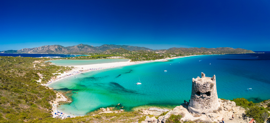 Panorama-Blick auf die Bucht von Villasimius auf Sardinien mit türkisfarbenem Wasser, einem weißen Sandstrand und einem alten, steinernen Wachturm im Vordergrund.