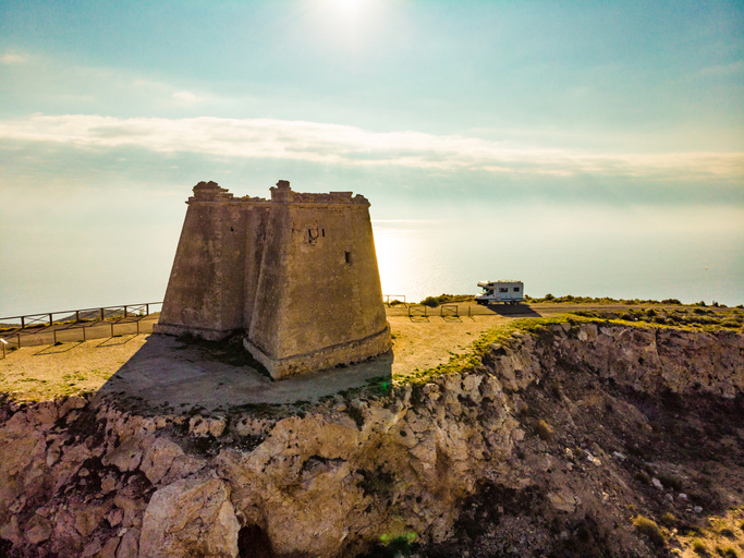 Ein alter, quadratischer Steinturm thront auf einer hohen Klippe über dem glitzernden Meer, daneben parkt ein Wohnmobil.
