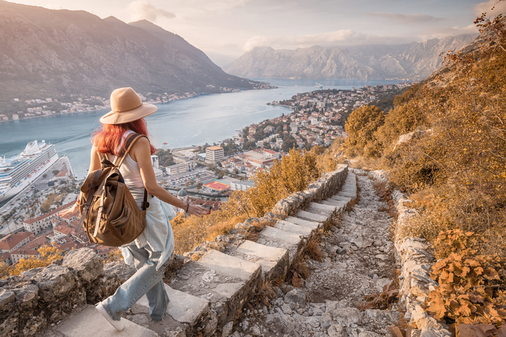 Eine Wanderin blickt von einem felsigen Bergpfad hinunter auf die mittelalterliche Stadt Kotor und den tiefblauen Fjord in Montenegro.