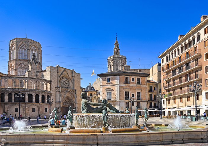 Ein großer Brunnen mit Skulpturen auf einem Platz in Valencia, dahinter ragen gotische Kirchtürme auf.