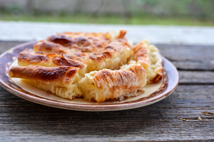 Ein goldbraun gebackener, aufgerollter Burek liegt in Stücke geschnitten auf einem rustikalen Teller.
