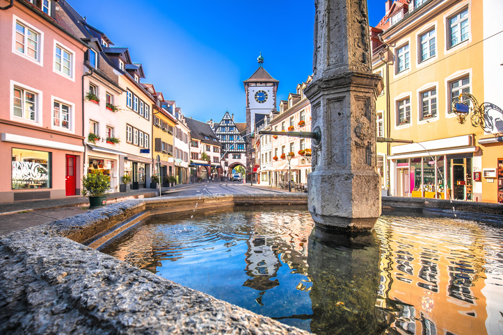 Ein Blick von einem Brunnen in der Fußgängerzone durch eine historische Gasse auf das weiße Martinstor mit seiner blauen Uhr.
