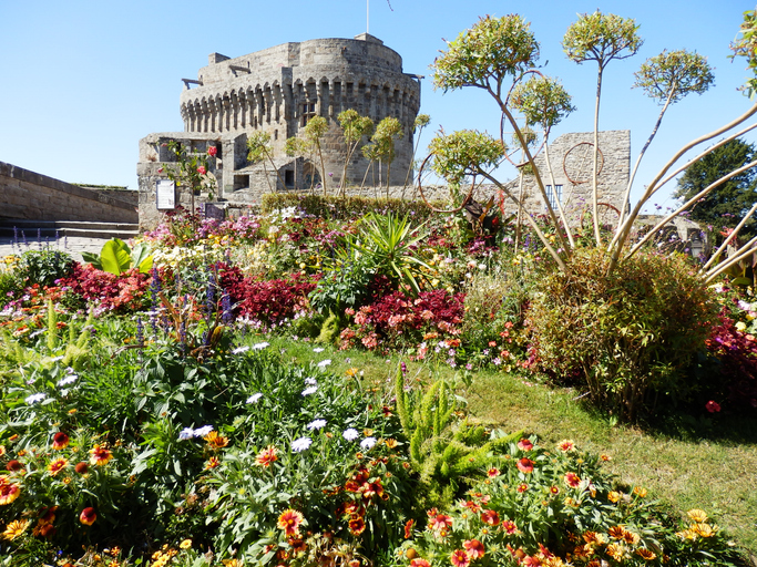 Ein buntes Blumenmeer im Vordergrund mit gelben, roten und weißen Blüten, dahinter ragt ein massiver runder Steinturm in den blauen Himmel.