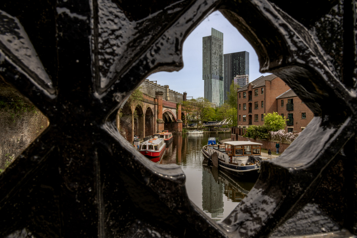 Durch die ornamentale Öffnung eines schwarzen Eisentores sieht man Hausboote auf einem Kanal und moderne Wolkenkratzer in der Ferne.
