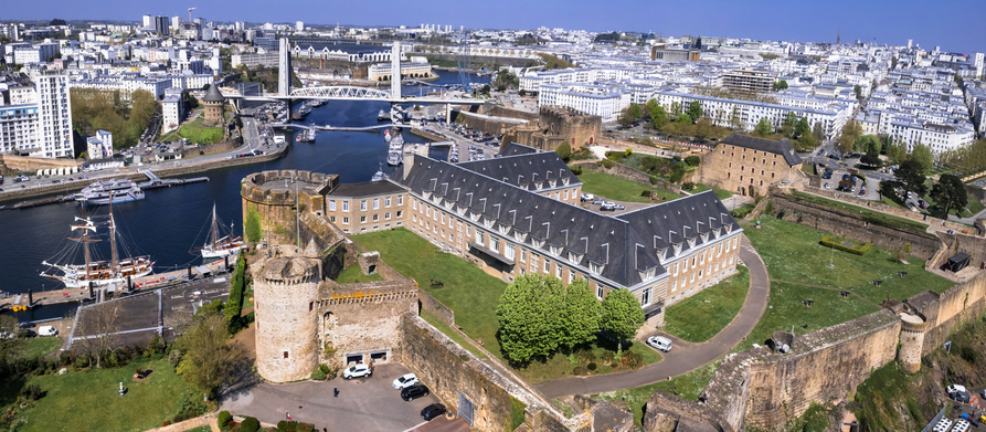 Luftaufnahme des mittelalterlichen Schlosses (Château de Brest) am Fluss Penfeld, mit modernen Hafenanlagen und Schiffen im Hintergrund.