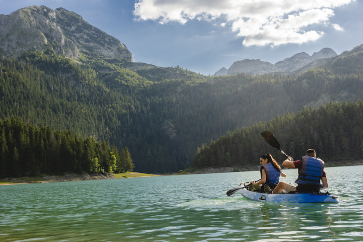 Ein Paar paddelt in einem blauen Kajak über einen klaren Bergsee im Durmitor-Nationalpark vor bewaldeten Gipfeln.