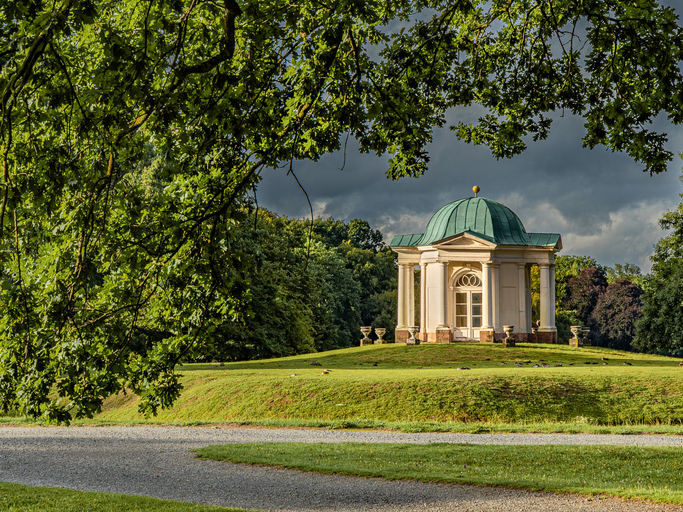 Ein kleiner, weißer Rundtempel mit grüner Kuppel auf einem sanften Grashügel, eingerahmt von alten Eichen.