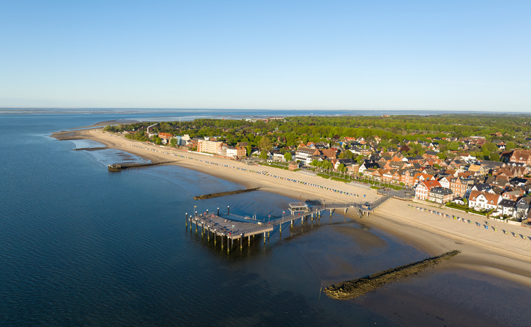 Luftaufnahme der Küstenlinie von Föhr mit einer Seebrücke, einem langen Sandstrand und dem Ort Wyk im Hintergrund.