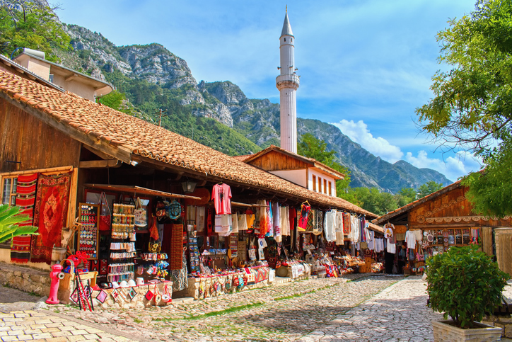 Ein gepflasterter Marktweg in der albanischen Stadt Kruja mit Holzhütten, bunten Teppichen und einem Minarett im Hintergrund.