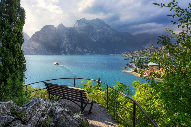 Eine hölzerne Parkbank auf einer Aussichtsplattform bietet einen perfekten Blick über den sonnigen Gardasee.