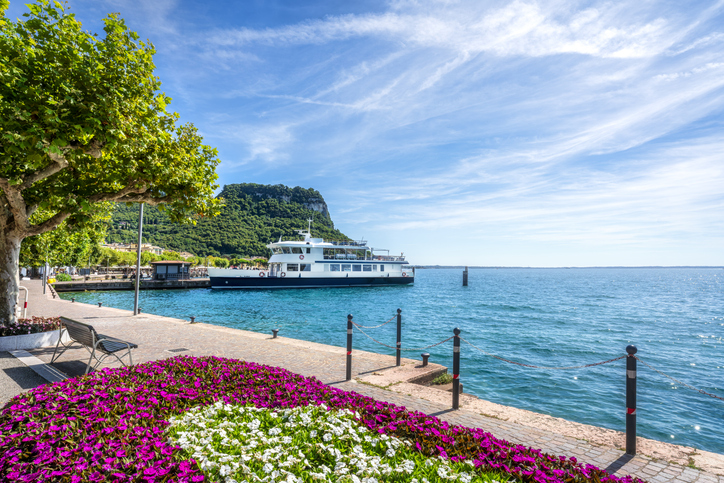 Ein Ausflugsschiff liegt in einem Hafen am Gardasee, davor blüht ein Blumenbeet in kräftigem Pink und Weiß.