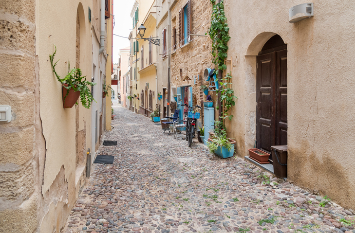 Eine enge, kopfsteingepflasterte Gasse in Alghero mit historischen Häusern, Pflanzen und einer alten Holztür