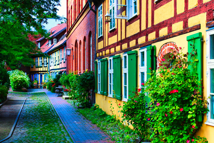 Eine schmale, mit Kopfsteinpflaster ausgelegte Gasse, gesäumt von gelben Fachwerkhäusern mit leuchtend grünen Fensterläden und Blumen.