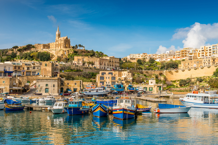 Bunte traditionelle Fischerboote schaukeln im Hafen von Mġarr auf Gozo vor einer Hügelstadt mit Kirche.