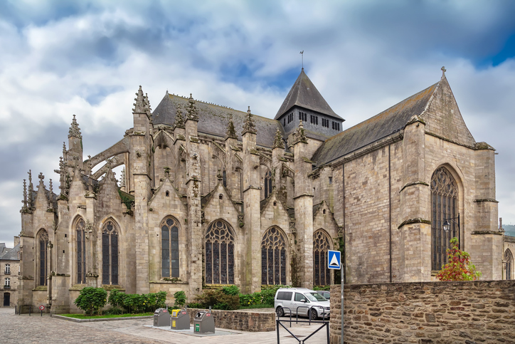 Die Außenansicht einer großen gotischen Kathedrale aus hellem Stein mit hohen Strebepfeilern und einem spitzen Turm unter wolkenverhangenem Himmel.
