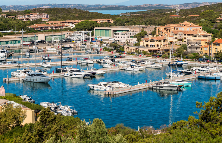 Blick auf den Yachthafen von Porto Cervo mit zahlreichen Luxusyachten vor der Kulisse pastellfarbener Gebäude bei sonnigem Wetter.